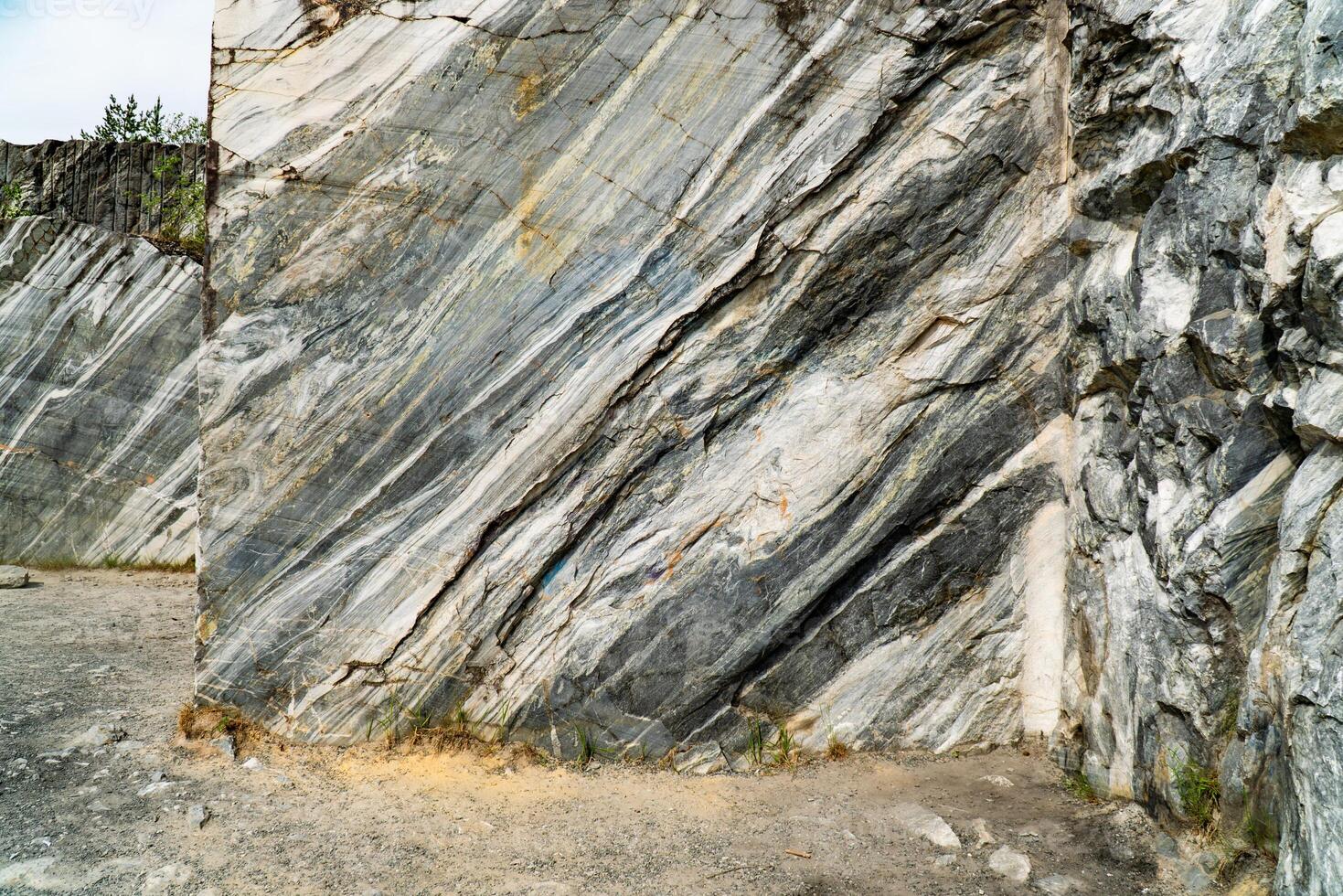 Scenic view of a marble quarry with distinct striations and pine trees in the background. The image highlights the contrast between the natural stone formations and the greenery under a bright blue sky. photo