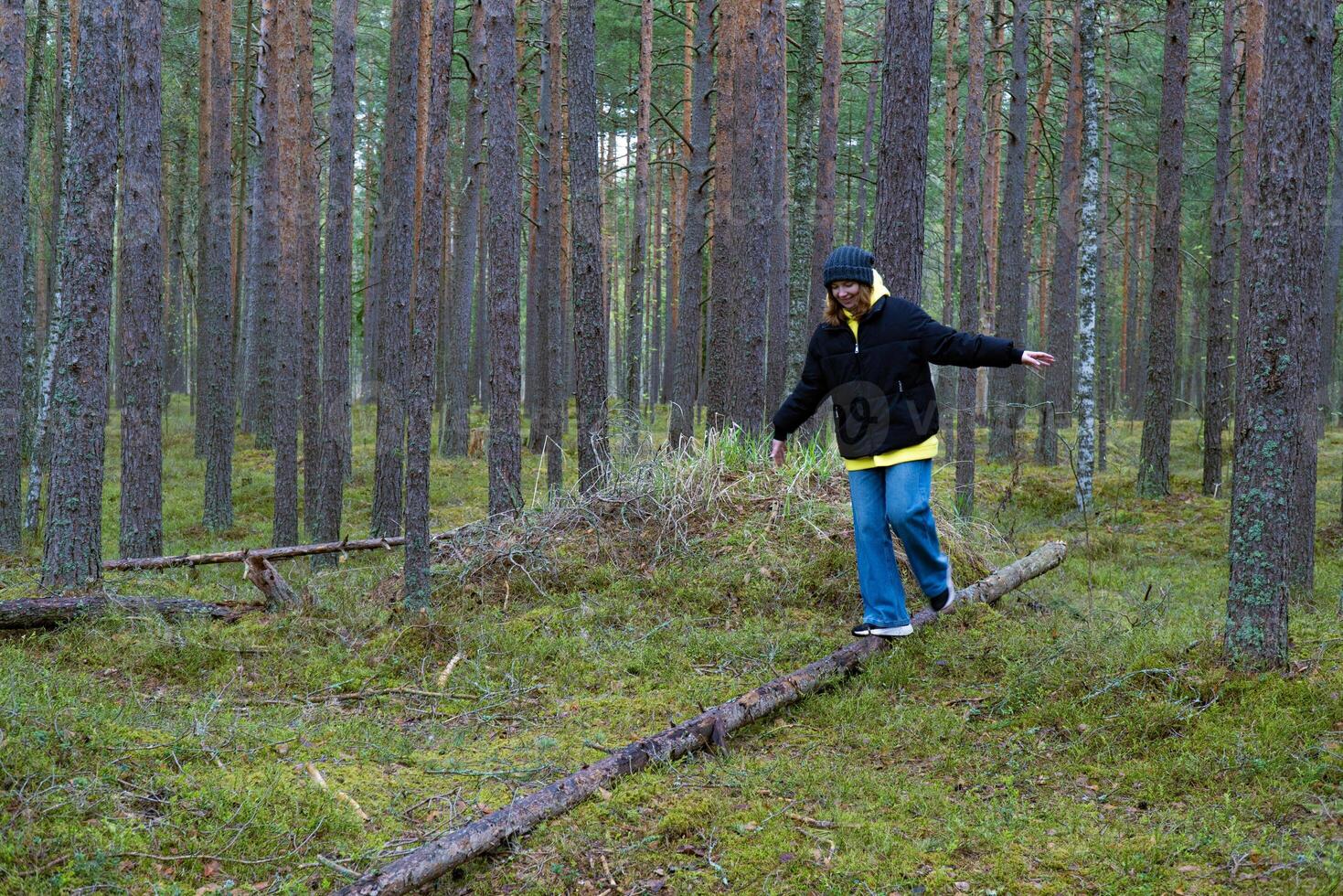 Image of a woman balancing on a fallen log in a dense pine forest. The scene captures a moment of outdoor adventure and nature exploration, ideal for themes related to hiking, balance, and woodland scenery photo