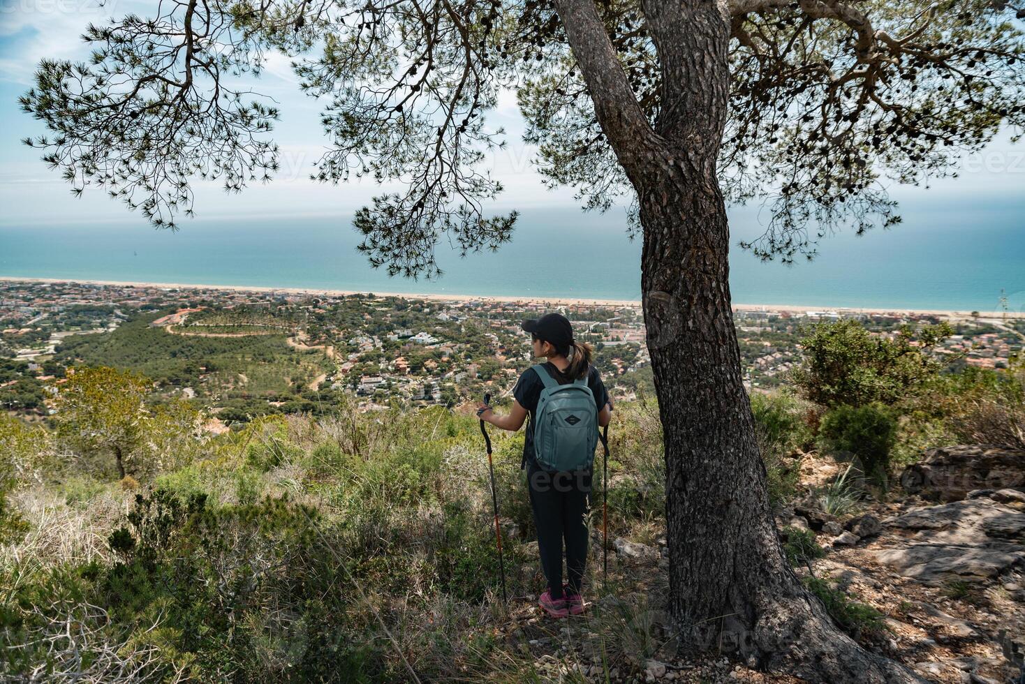Hiker with a backpack and mountain sticks, contemplates the Mediterranean coast from above and below a tree. photo