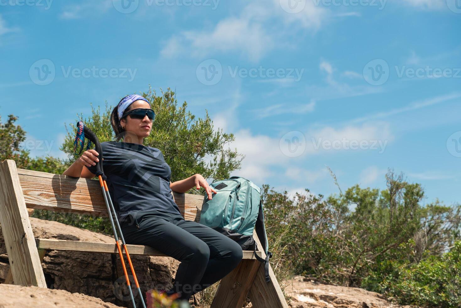 Woman with hiking sticks and backpack rests on a wooden bench located on top of a mountain. photo