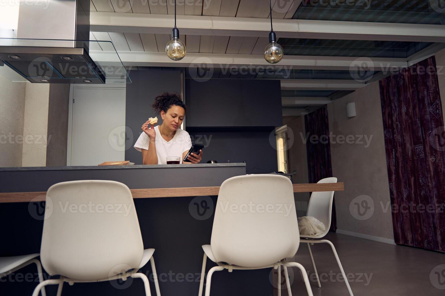 Beautiful multi ethnic woman 40s, using smartphone, checking social media content, browsing web, scrolling newsfeed, standing at kitchen counter with a sandwich in hand, enjoying her breakfast at home photo