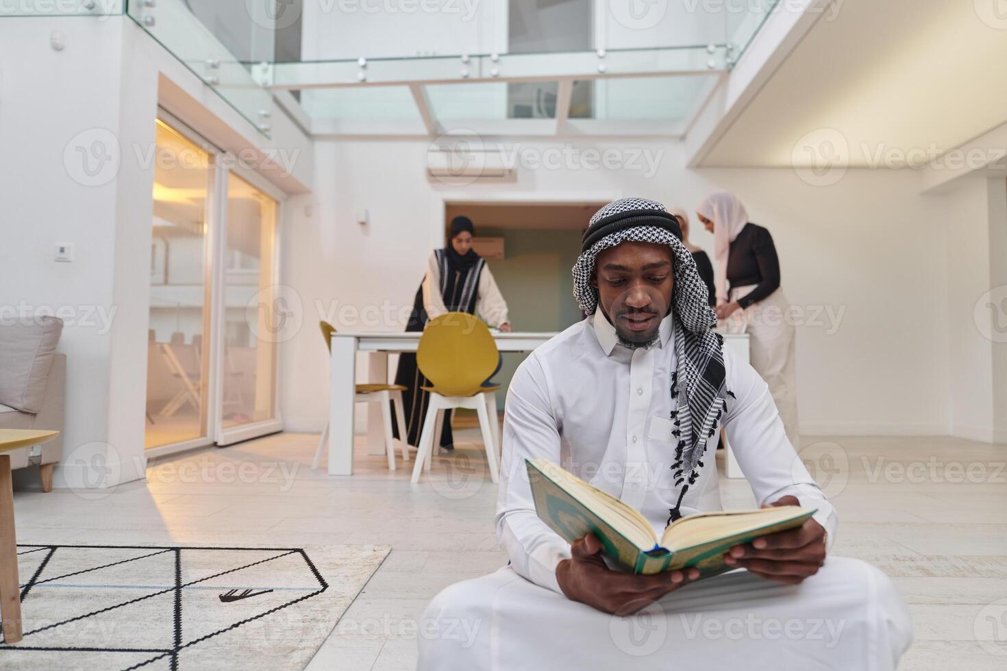 In the sacred month of Ramadan, an African American Muslim man engrossed in reading the Holy Quran is surrounded by a backdrop where three hijab-wearing women prepare and serve food for iftar photo