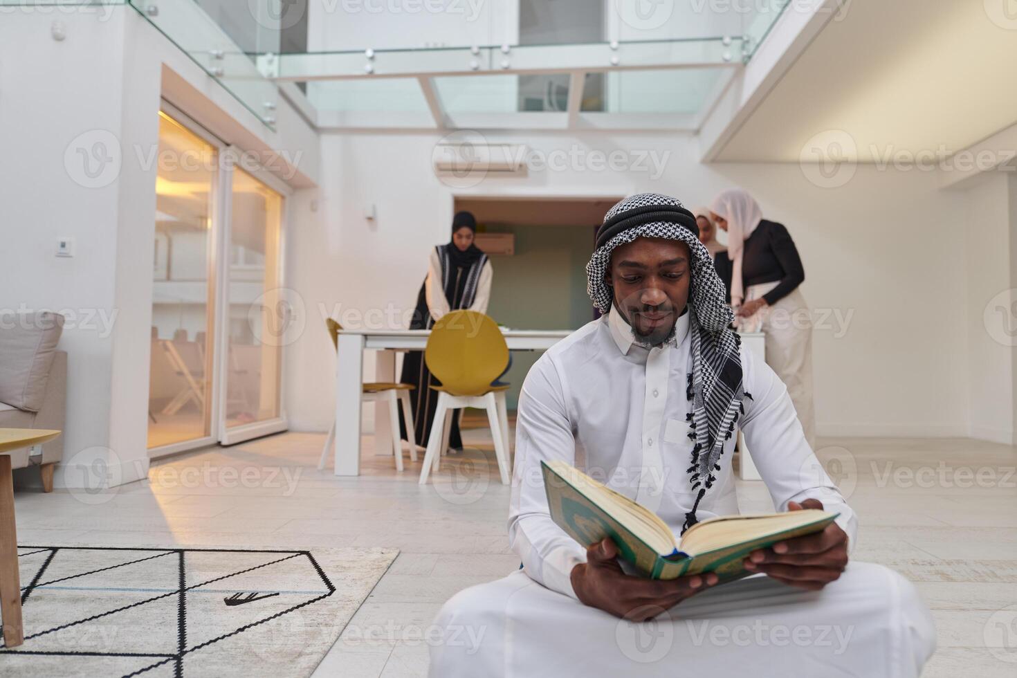 In the sacred month of Ramadan, an African American Muslim man engrossed in reading the Holy Quran is surrounded by a backdrop where three hijab-wearing women prepare and serve food for iftar photo