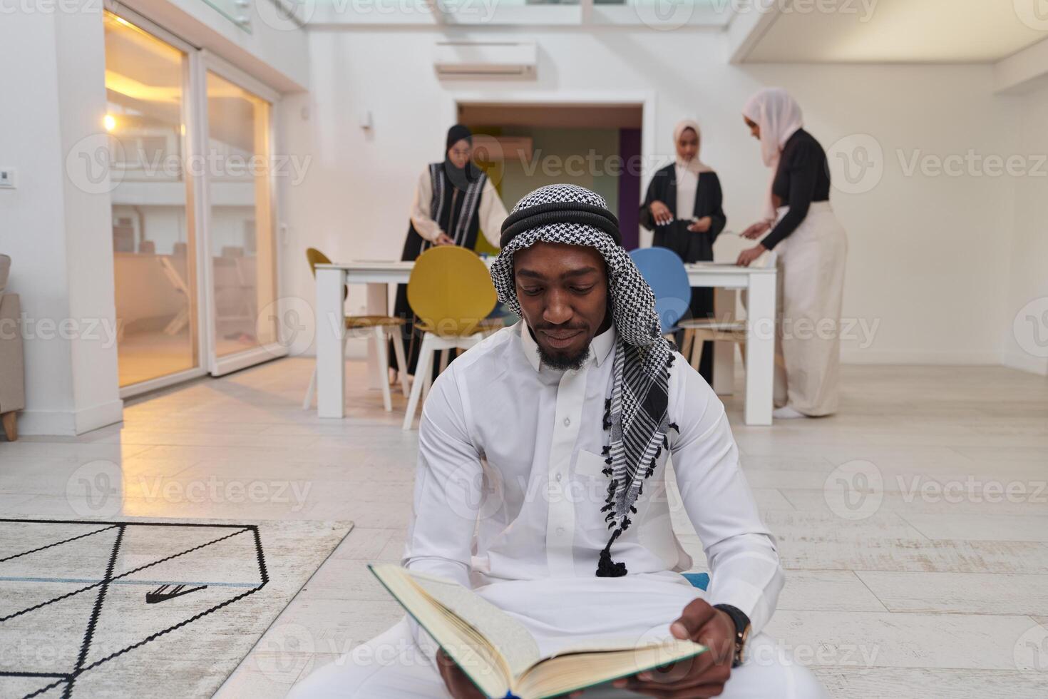 In the sacred month of Ramadan, an African American Muslim man engrossed in reading the Holy Quran is surrounded by a backdrop where three hijab-wearing women prepare and serve food for iftar photo