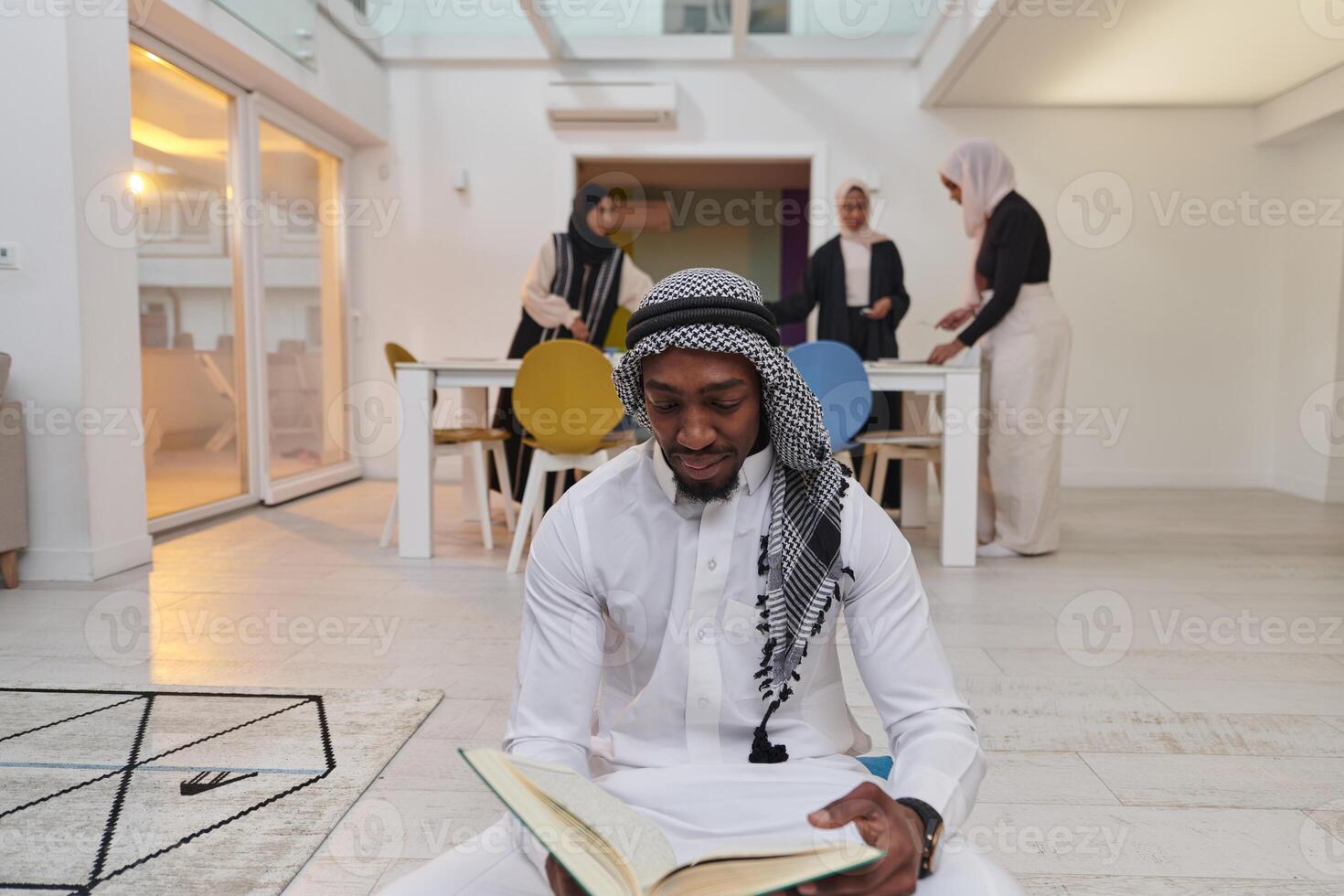 In the sacred month of Ramadan, an African American Muslim man engrossed in reading the Holy Quran is surrounded by a backdrop where three hijab-wearing women prepare and serve food for iftar photo