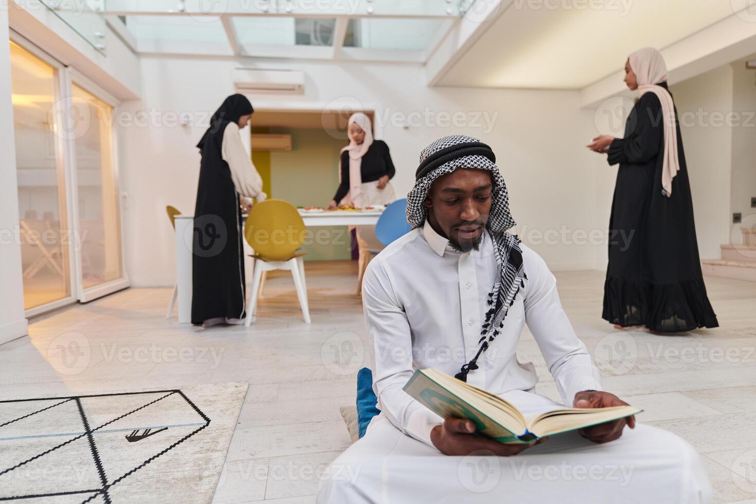 In the sacred month of Ramadan, an African American Muslim man engrossed in reading the Holy Quran is surrounded by a backdrop where three hijab-wearing women prepare and serve food for iftar photo