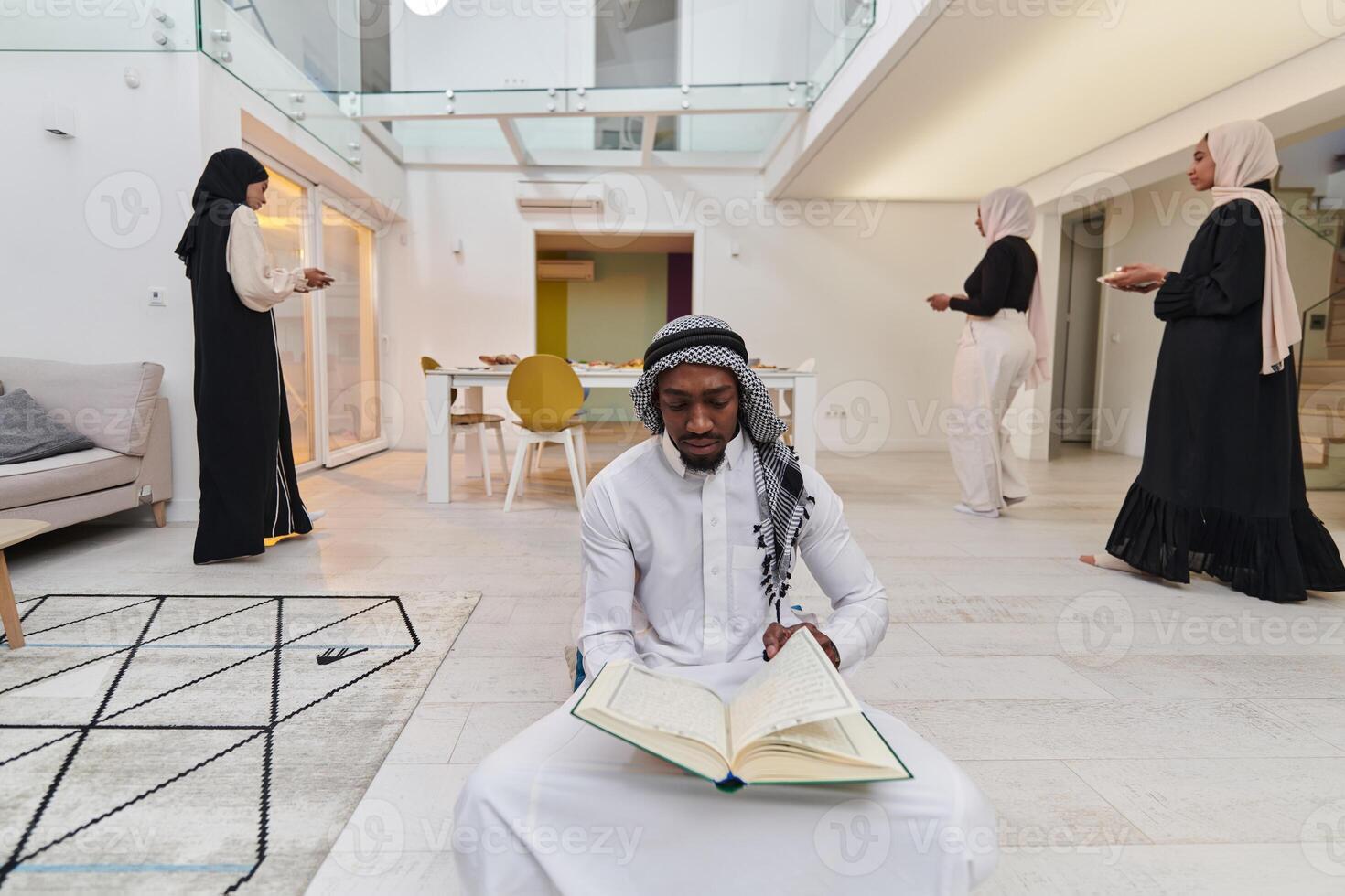 In the sacred month of Ramadan, an African American Muslim man engrossed in reading the Holy Quran is surrounded by a backdrop where three hijab-wearing women prepare and serve food for iftar photo