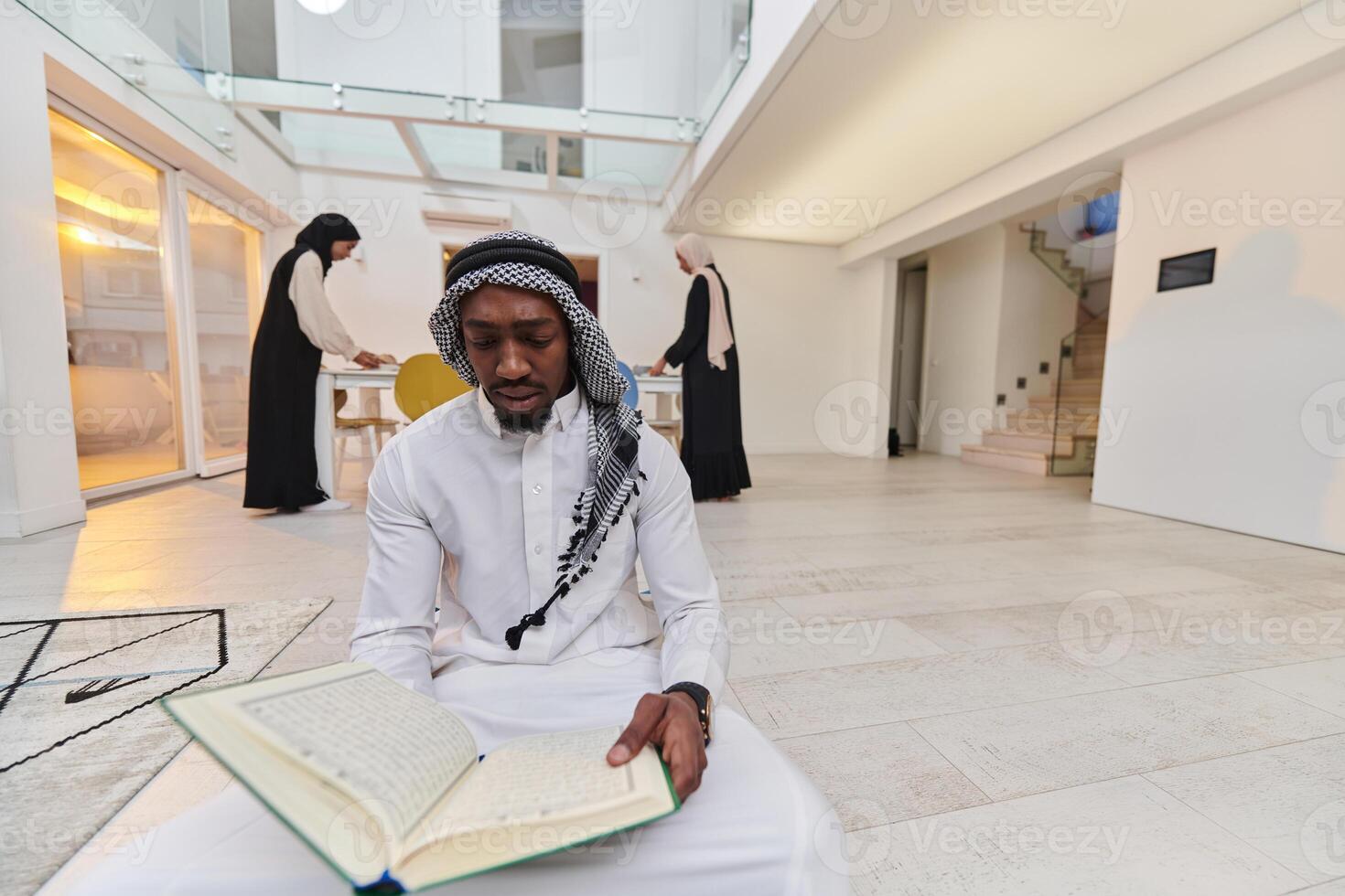 In the sacred month of Ramadan, an African American Muslim man engrossed in reading the Holy Quran is surrounded by a backdrop where three hijab-wearing women prepare and serve food for iftar photo