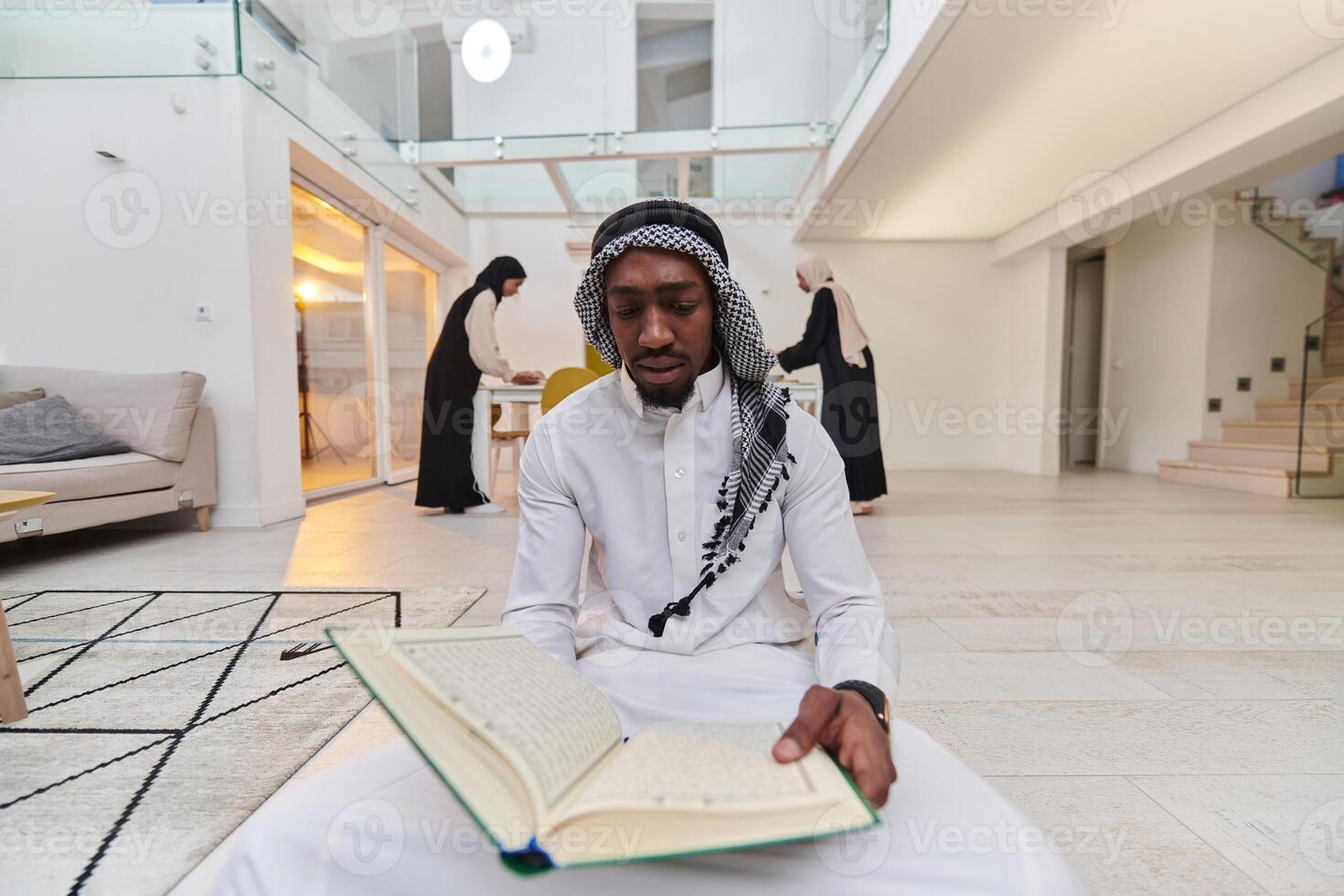 In the sacred month of Ramadan, an African American Muslim man engrossed in reading the Holy Quran is surrounded by a backdrop where three hijab-wearing women prepare and serve food for iftar photo