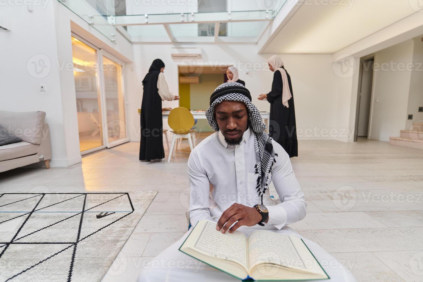 In the sacred month of Ramadan, an African American Muslim man engrossed in reading the Holy Quran is surrounded by a backdrop where three hijab-wearing women prepare and serve food for iftar photo