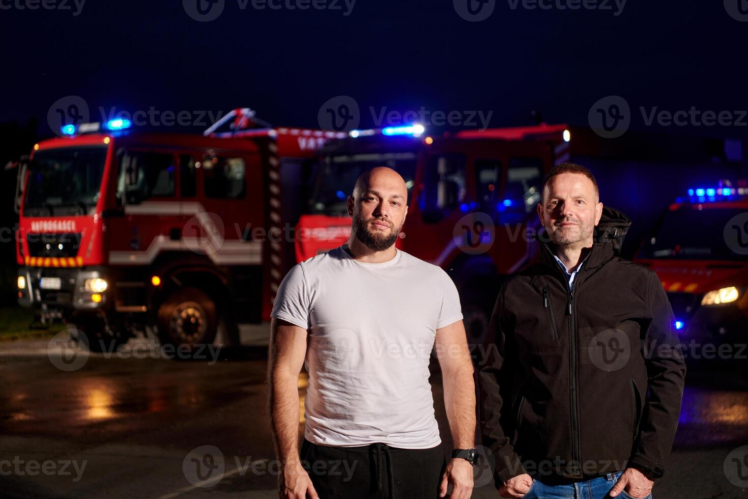 Group of firefighters, dressed in civilian clothing, stand in front of fire trucks during the night, showcasing a moment of camaraderie and unity among the team as they reflect on their duties and the photo