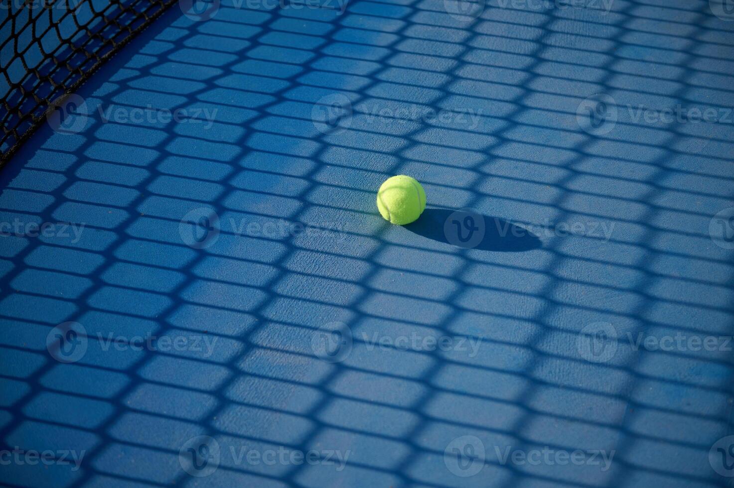 Tennis ball on blue court surface with net shadow in sunlight photo