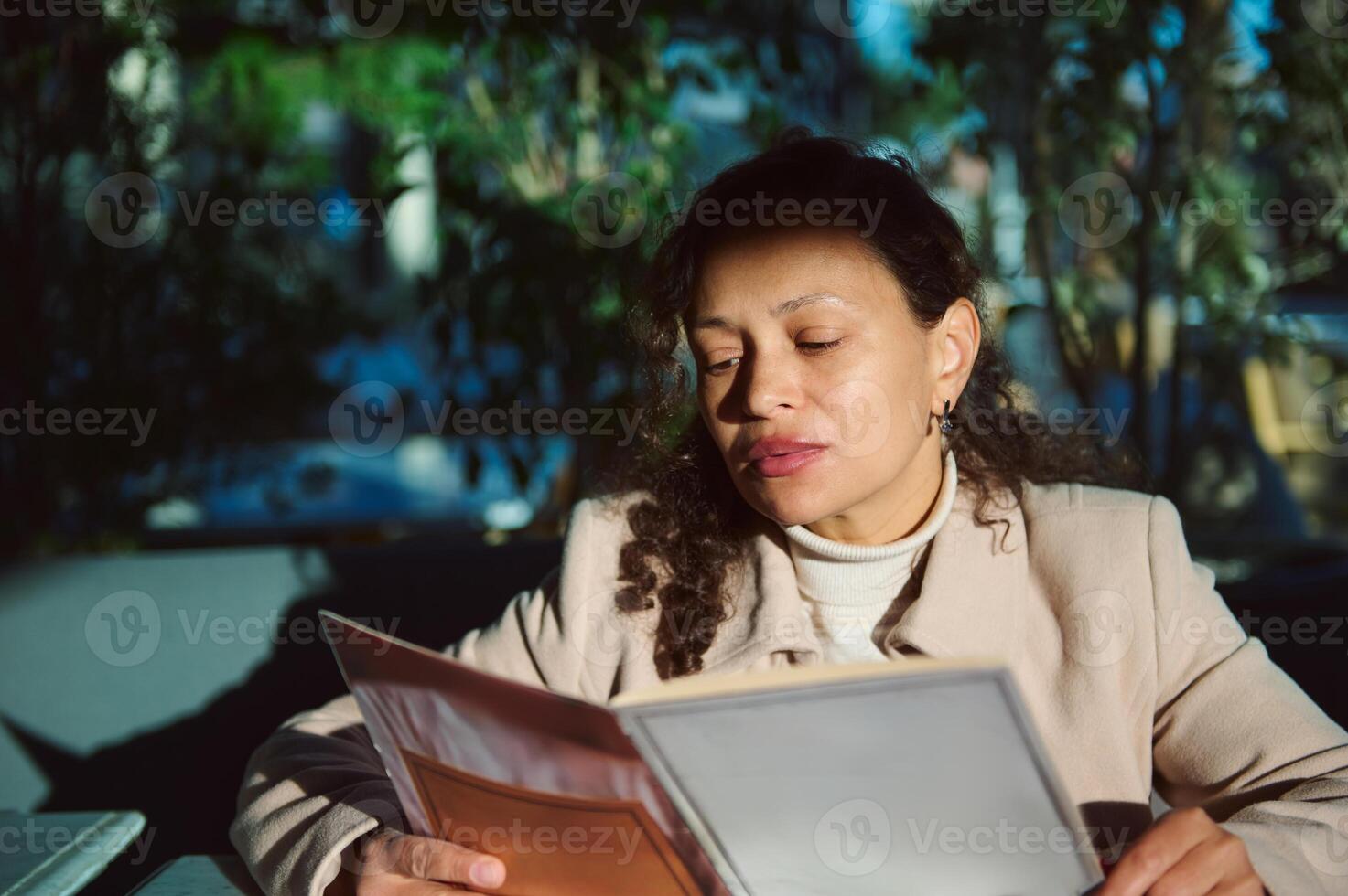 Woman reading a menu at an outdoor cafe in bright sunlight with greenery in the background photo