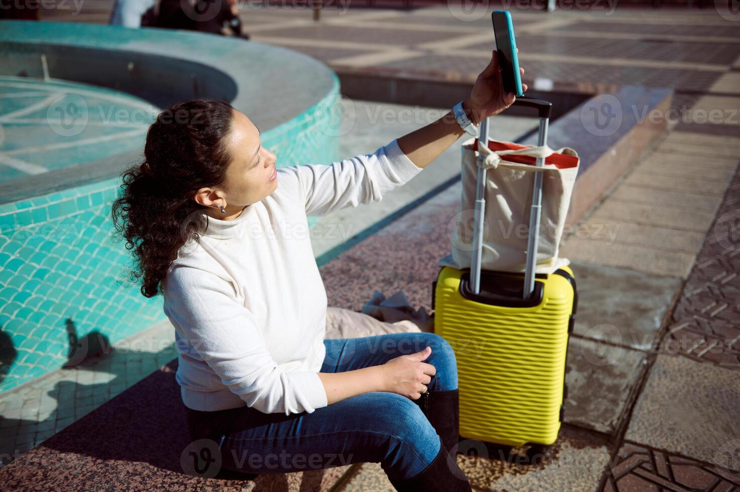 Woman traveler sitting by fountain taking selfie with smartphone and yellow luggage photo