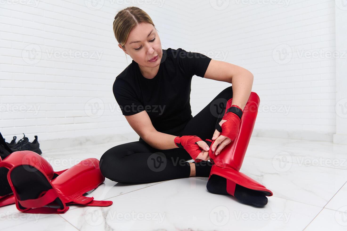 Beautiful determines athletic woman boxer fighter putting on red kickboxing leg pads guard. Sparring shin guards, isolated over white background. Martial art, combat, challenge. Kickboxing concept photo