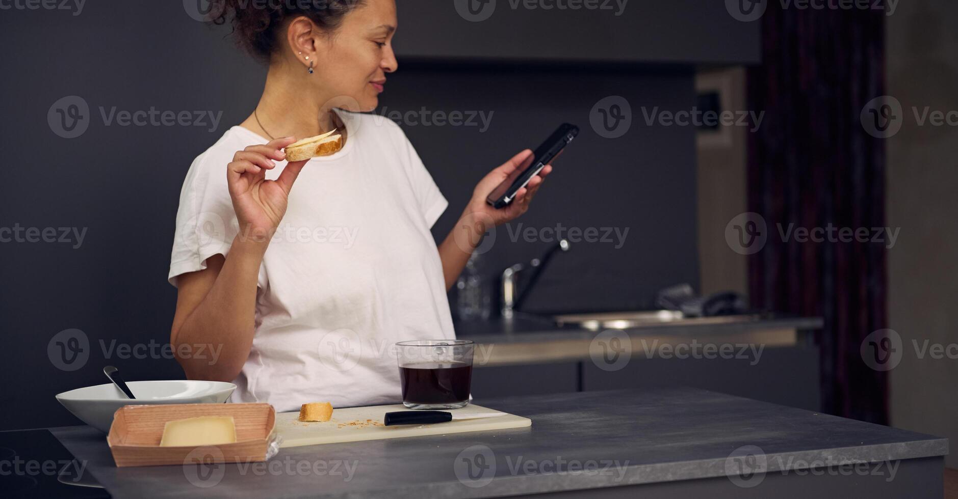 Beautiful multi ethnic woman 40s, using smartphone, checking social media content, browsing web, scrolling newsfeed, standing at kitchen counter with a sandwich in hand, enjoying her breakfast at home photo
