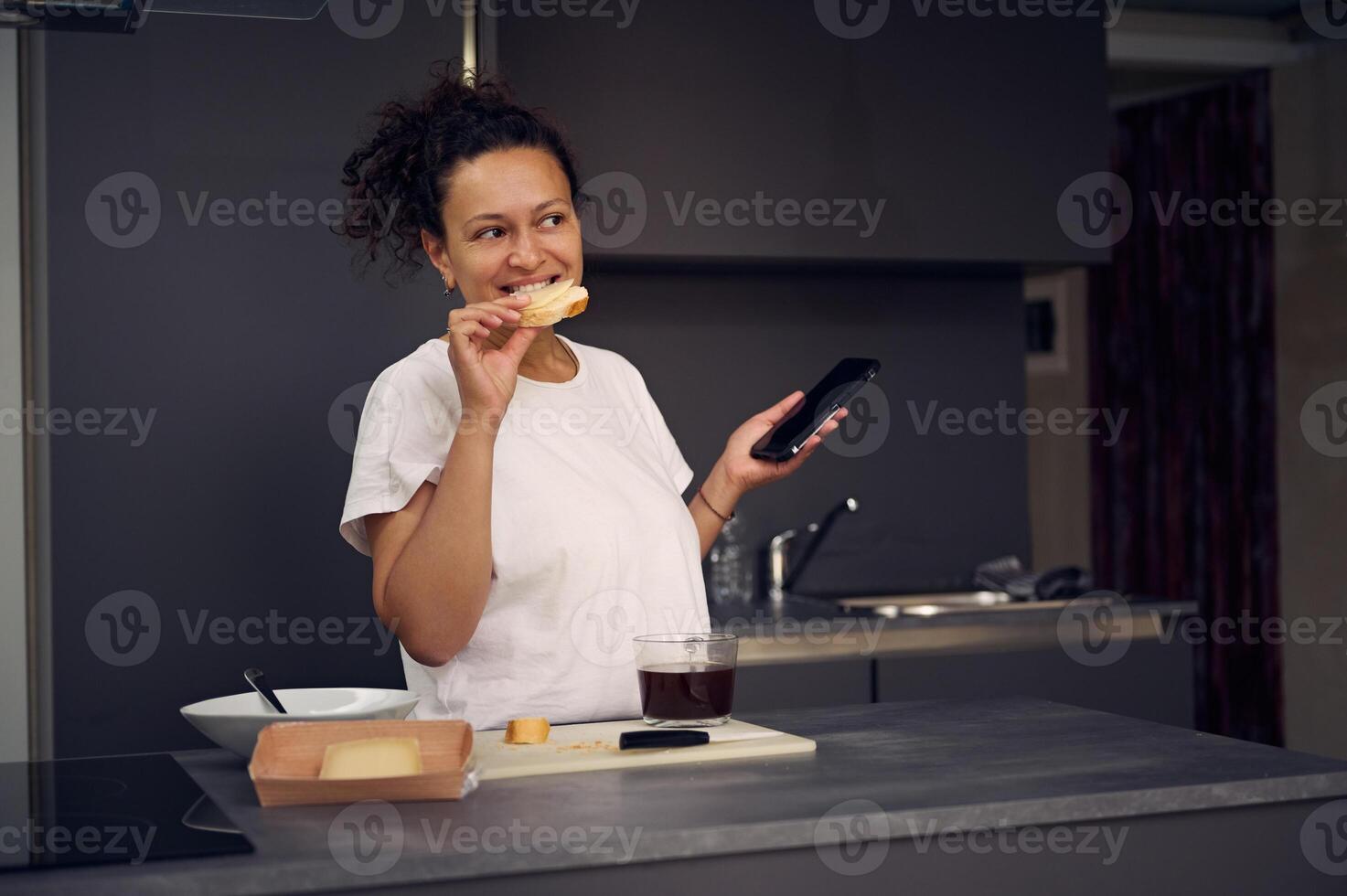 Beautiful multi ethnic woman 40s, using smartphone, checking social media content, browsing web, scrolling newsfeed, standing at kitchen counter with a sandwich in hand, enjoying her breakfast at home photo