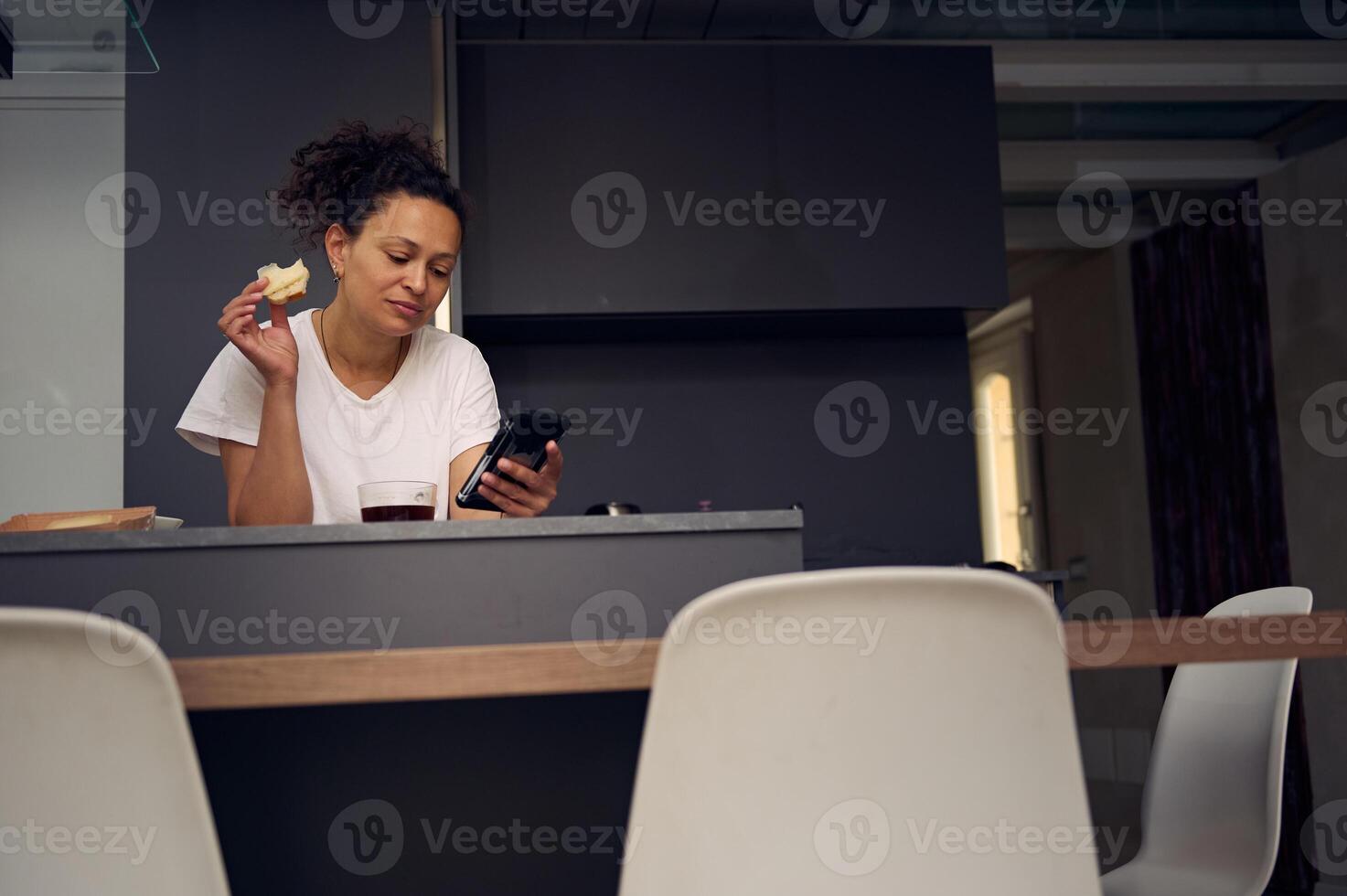 Confident mixed race woman 40s, using smartphone, checking social media content, browsing web, scrolling newsfeed, standing at kitchen counter with a sandwich in hand, enjoying her breakfast at home photo
