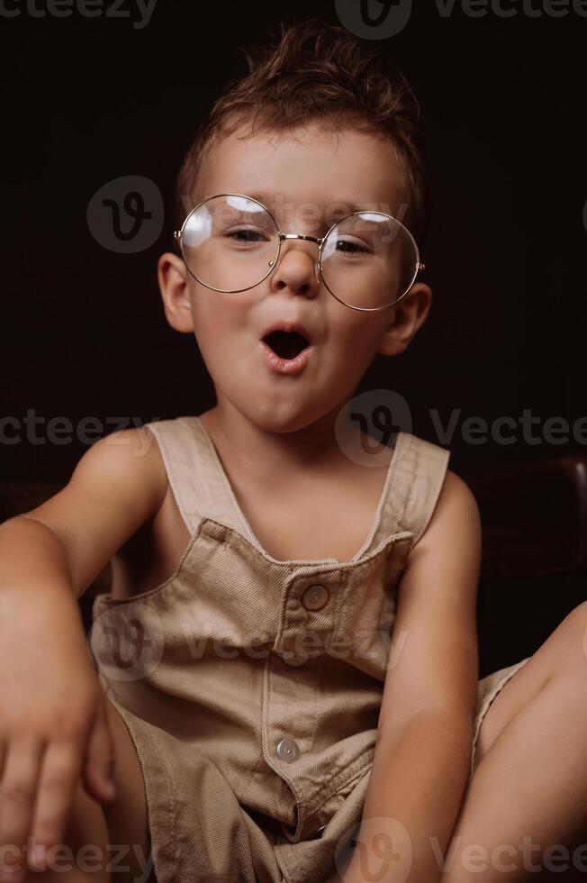 stylish Caucasian boy with glasses and a fashionable hairstyle in a jumpsuit is sitting on a wooden chair photo