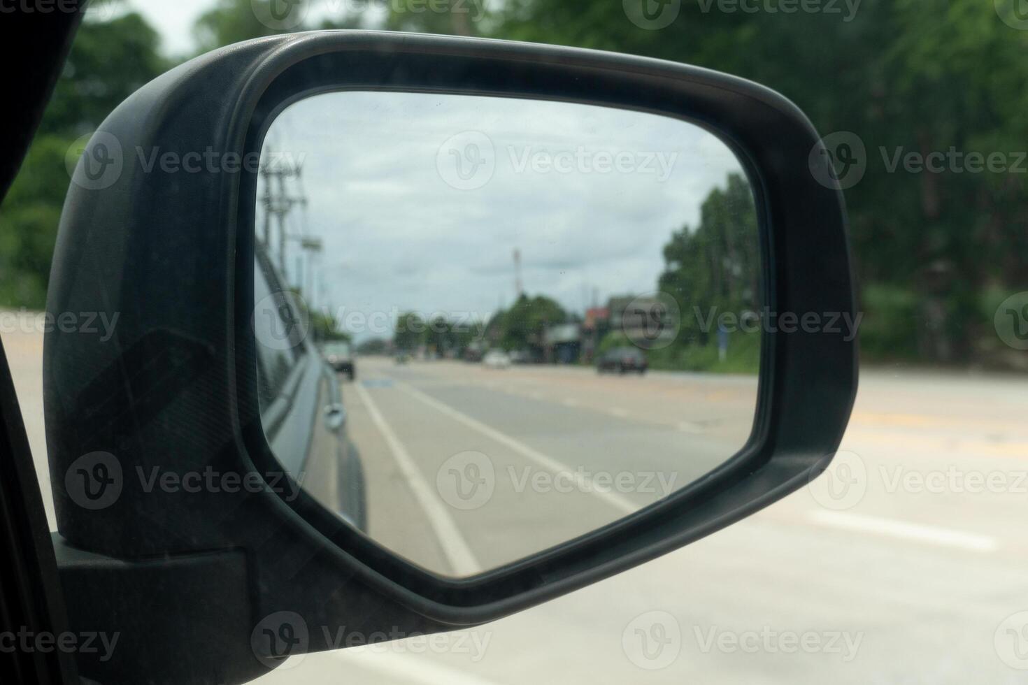 Inside view of mirrors wing. Rear view of a gray car with asphalt road and green trees with small city in the daytime. Clear traffic in rural areas. photo