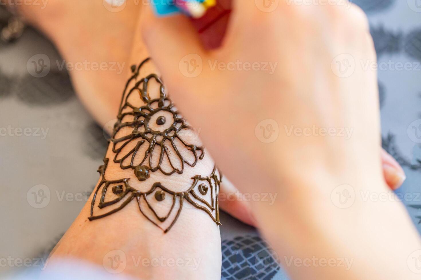 This image shows a woman applying a henna tattoo to another womans hand. The womans hand is resting on a table with a pineapple patterned tablecloth. The henna tattoo is a intricate design of flowers and swirls. photo