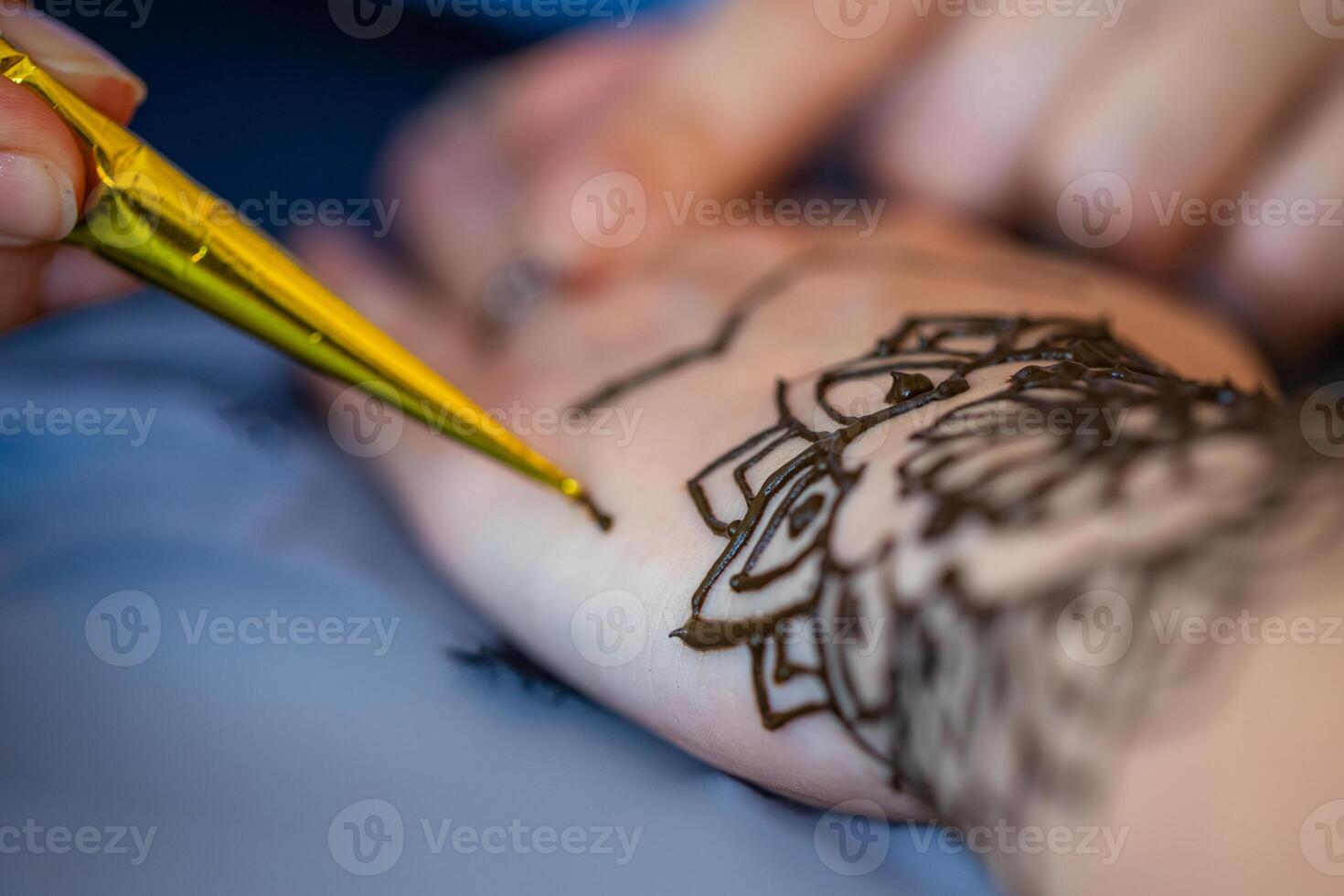 A close-up image of a woman receiving a henna tattoo on her wrist. The artist is using a cone filled with henna paste to meticulously draw an intricate design on the womans skin. The background is a fabric with a pattern of pineapples. photo