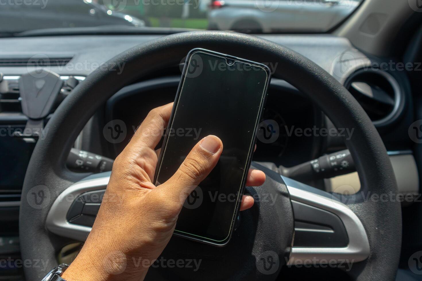 Male hand holding a smartphone with a car steering wheel in the background. Driving while using a smart phone concept. photo