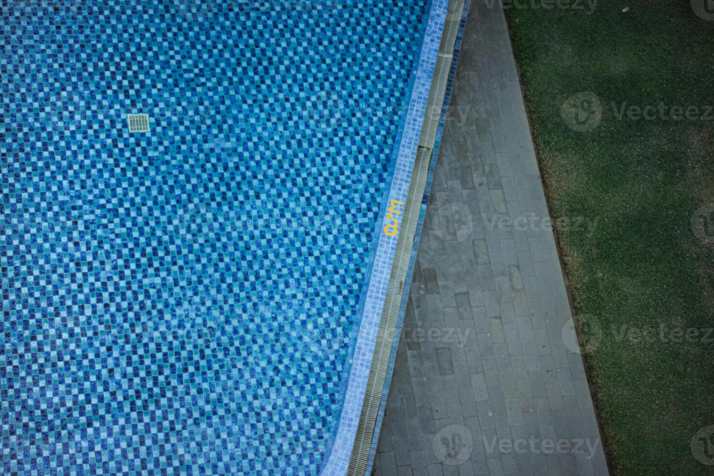 an aerial view of a swimming pool with a checkered tile pattern. The pool is surrounded by a concrete deck and a grassy area or plants or flower area. The scene is calm and inviting. photo