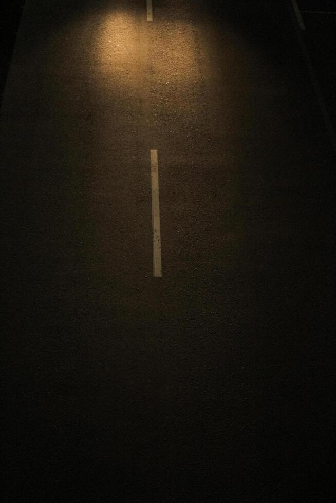 a dark asphalt road at night. A single white lane marker is visible in the center of the road, illuminated by a bright car light source. photo