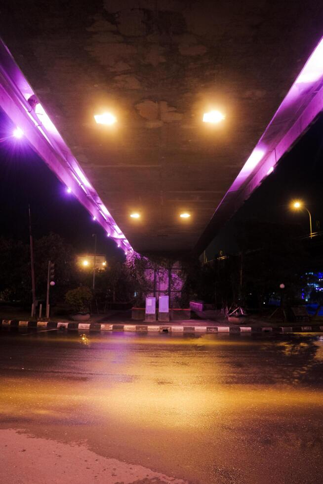 a view from underneath a bridge at night, with purple and yellow lights illuminating the scene. The wet pavement reflects the lights photo