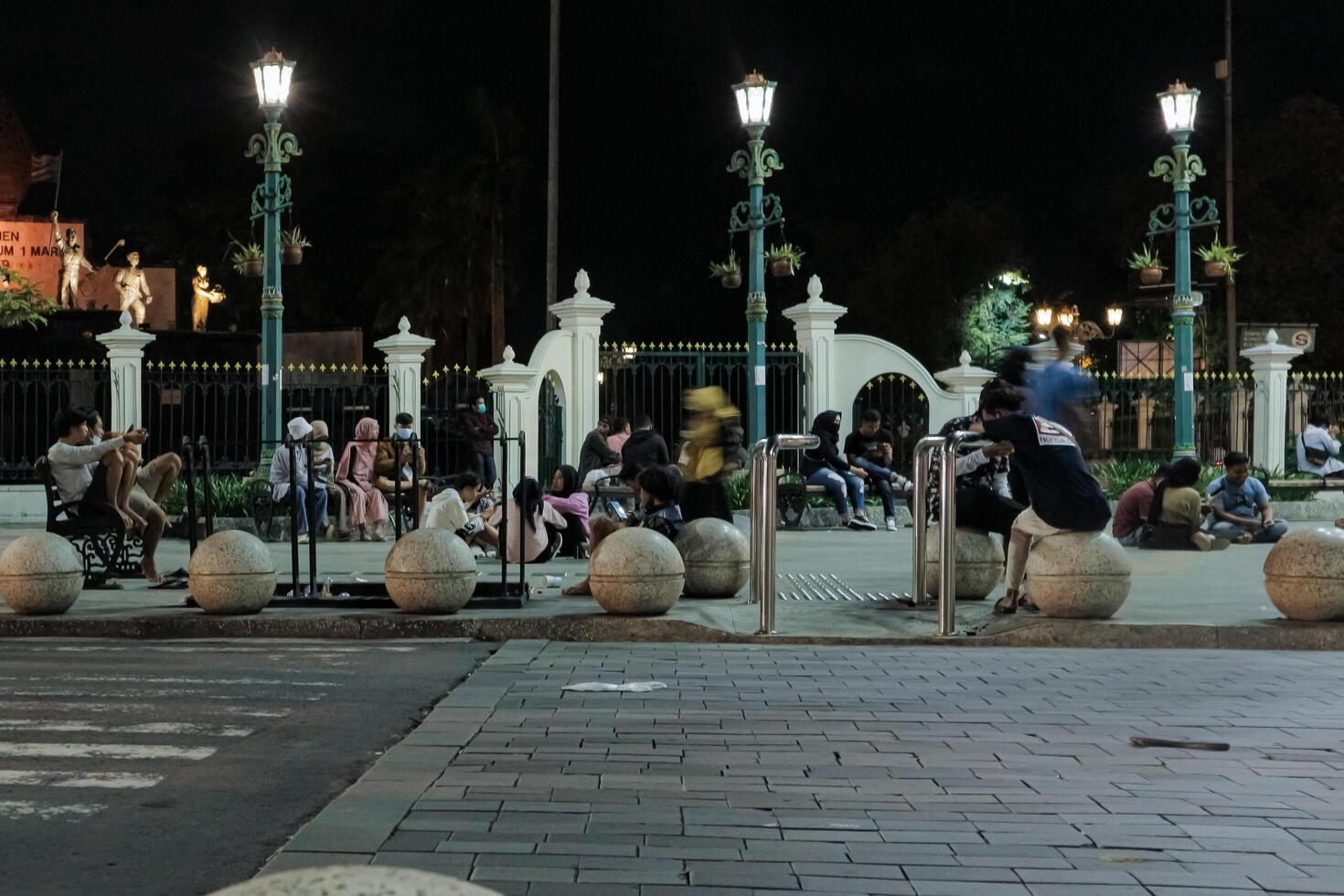 a group of people sitting on benches in kilometer nol, yogyakarta and round stone structures in a public square at night. The scene is peaceful and relaxed, with a sense of community and togetherness photo