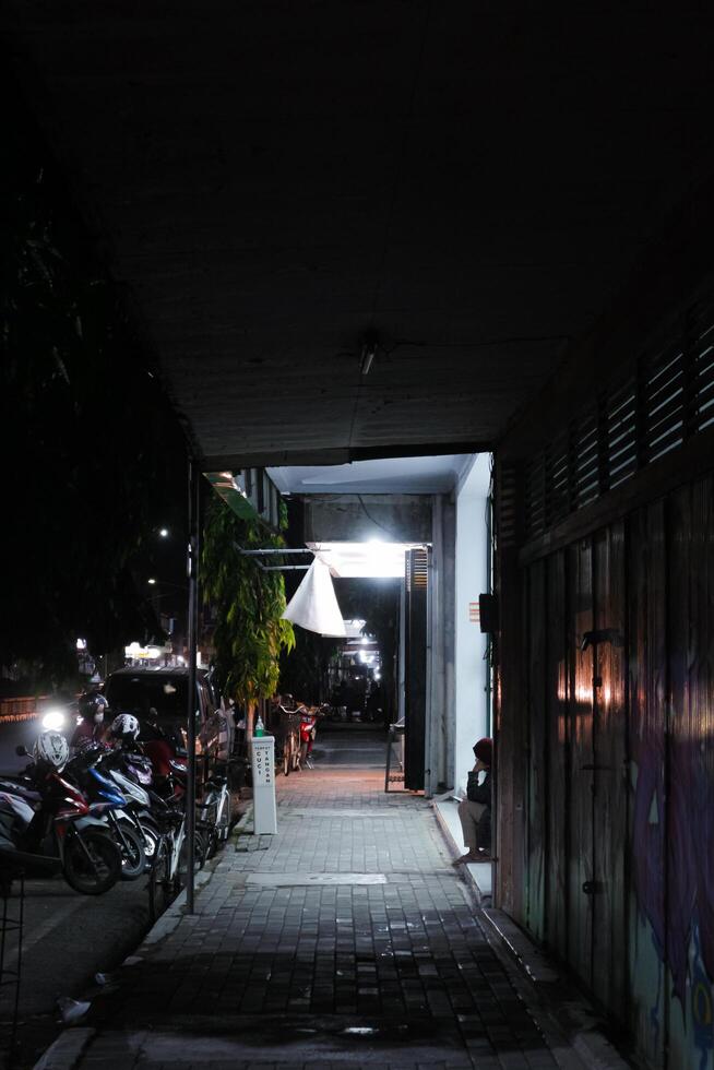 a narrow alleyway at night, with a person sitting on the edge of the sidewalk. The alleyway is dimly lit, and the shadows cast by the buildings and the person create a sense of mystery and intrigue photo