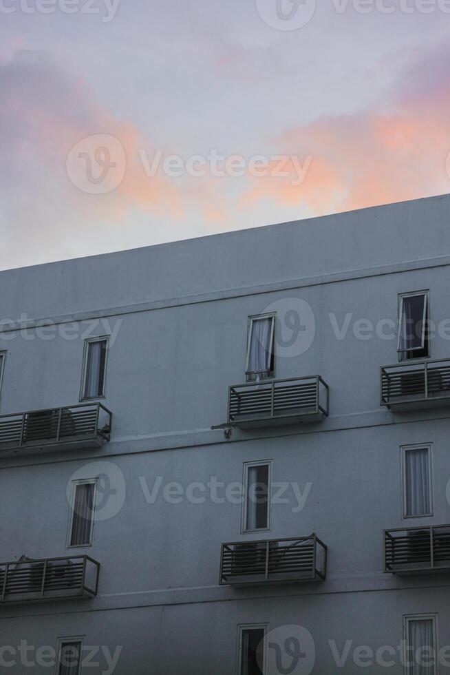 white apartment building with several windows and balconies. The sky is a soft pink and purple at sunset, suggesting a beautiful sunset. The scene is peaceful and serene photo
