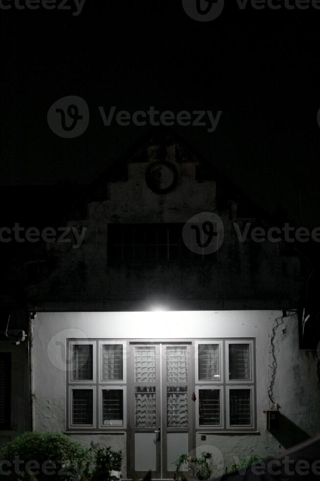 dimly lit house at night, with a single light illuminating the front door. The house has a classic design with a gabled roof and a circular window. photo
