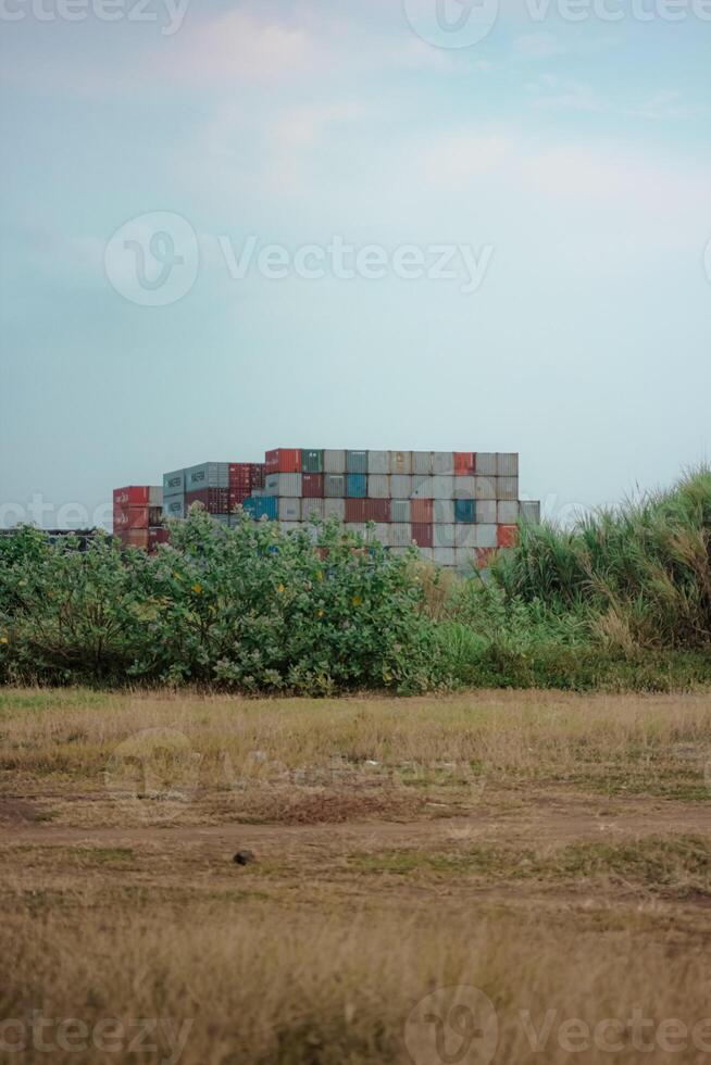a stack of shipping containers in the distance, partially obscured by a row of green bushes. a field of dry grass, with a dirt path running through it. sky is a pale blue with a few wispy clouds. photo