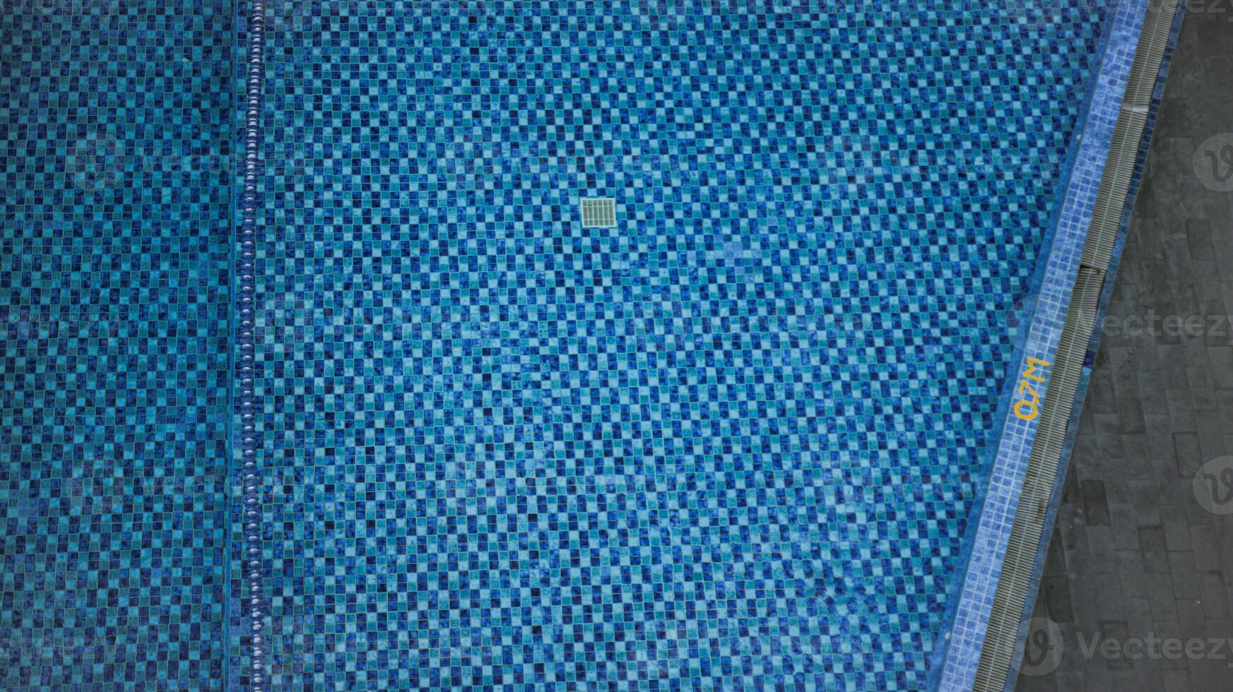 an aerial view of a swimming pool with a checkered tile pattern. The pool is surrounded by a concrete deck and a grassy area or plants or flower area. The scene is calm and inviting. photo