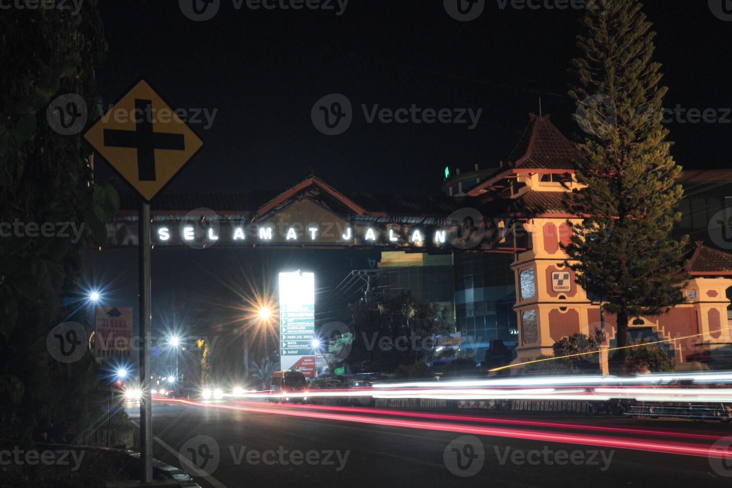 A vibrant nighttime scene at the entrance to the town of Pemalang, featuring a welcoming archway with a Selamat Jalan sign, illuminated by bright lights. Cars whiz across road, with streaks lights. photo