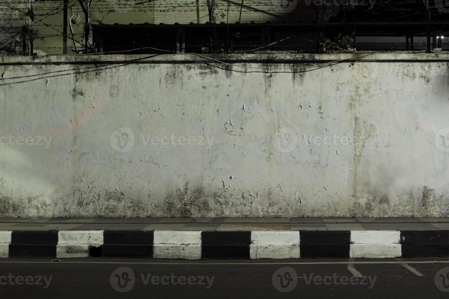 concrete wall with a black and white curb in front of it. The wall is weathered and cracked, and there is a barbed wire fence on top of it. The scene is dark and desolate photo