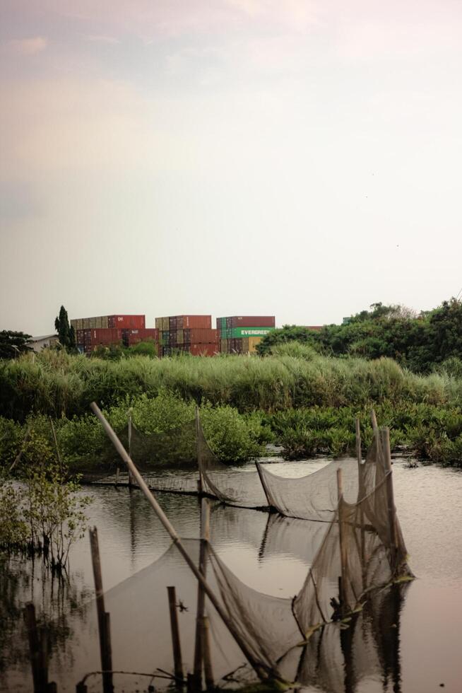 Serene wetland landscape with suspended fishing nets and colorful cargo containers in the distance. The tranquil, reflective waters and lush vegetation create a harmonious, eco-industrial scene. photo