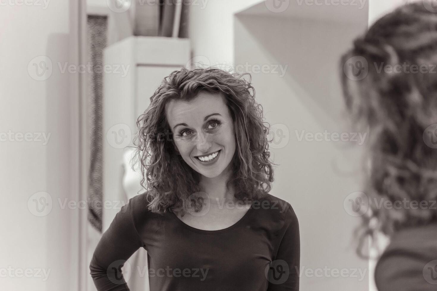 Curly-haired woman in a black top smiling at her reflection in a mirror, expressing satisfaction photo