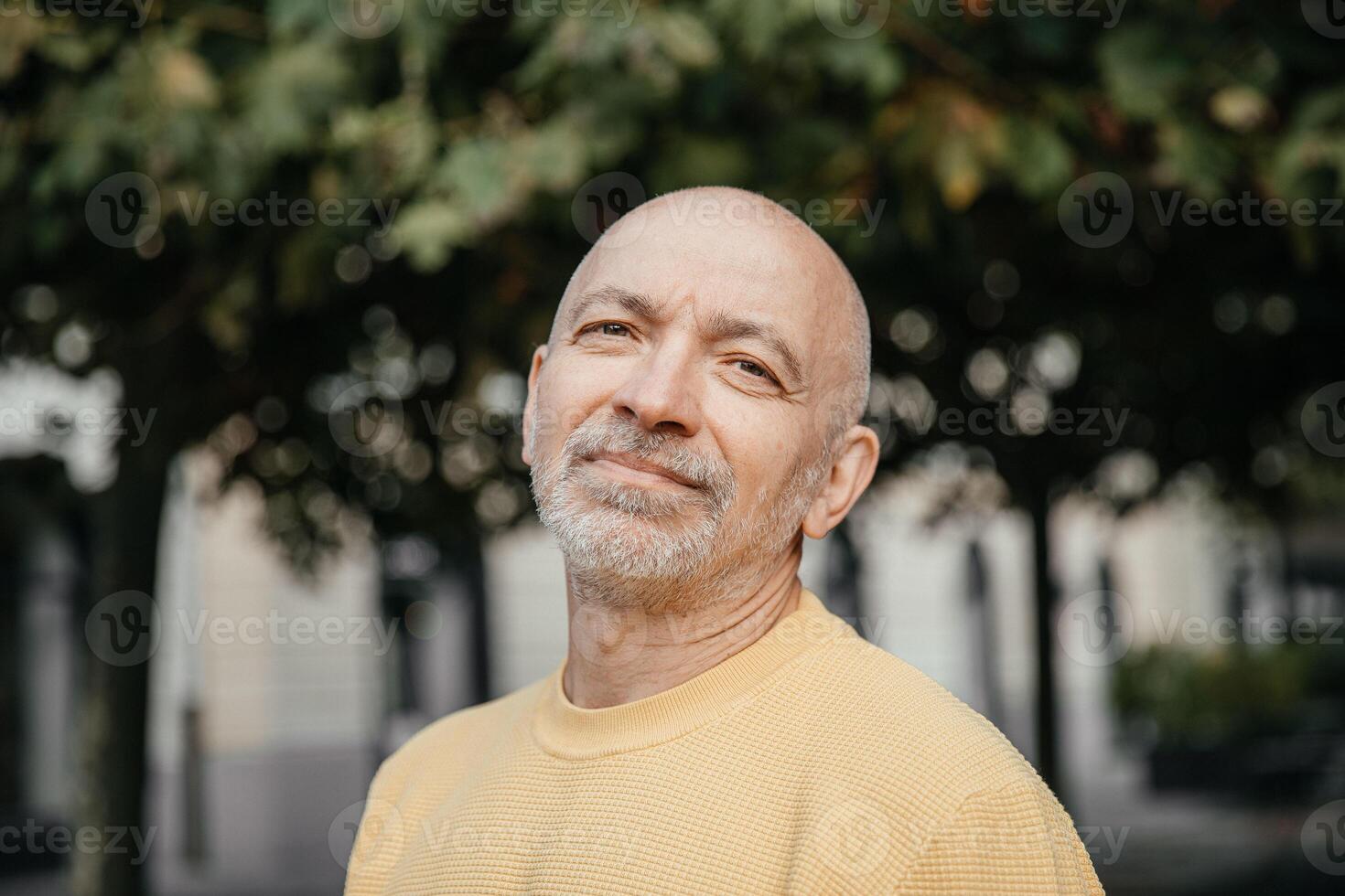 Senior man with a gentle smile, wearing a yellow sweater, standing under the shade of trees in an urban environment photo