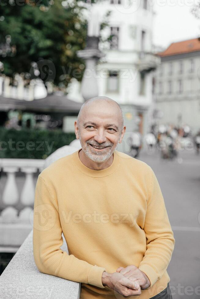 Cheerful bald senior man in a yellow sweater, leaning on a stone balustrade in an urban environment, radiating happiness photo