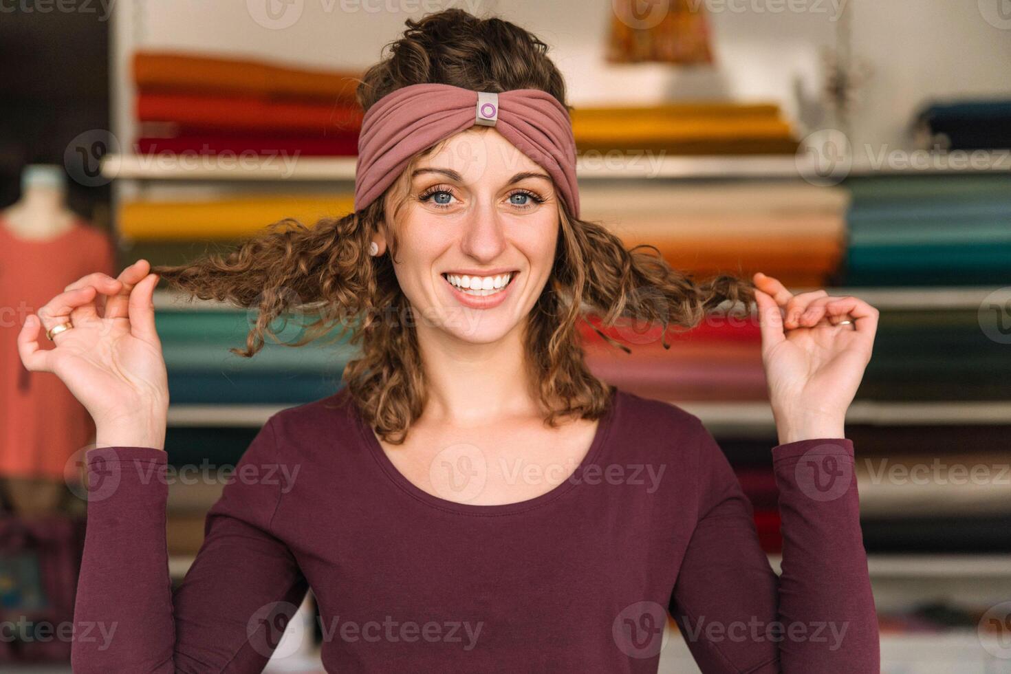 A playful fashion designer with curly hair and sparkling eyes showcases a mauve headband, standing in front of colorful fabric rolls in her workshop photo