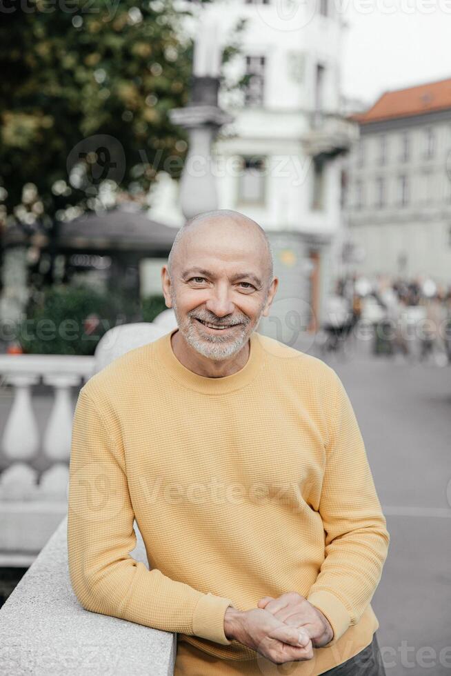 Smiling Senior Man in Yellow Sweater Leaning on Urban Balustrade photo