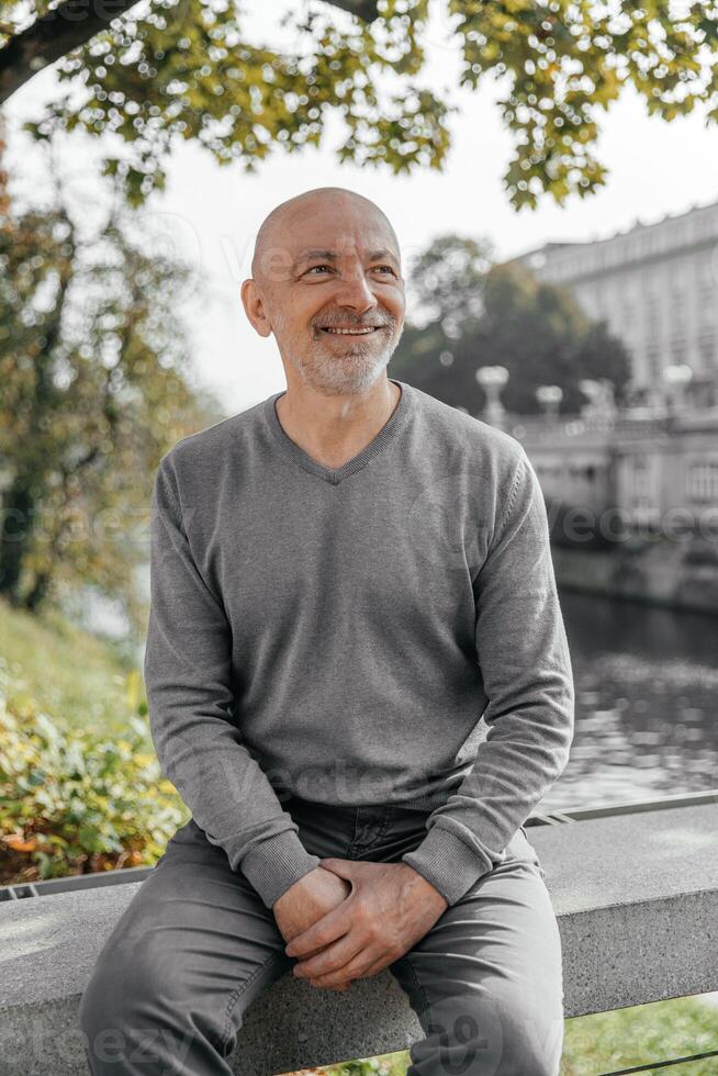 Elderly man sitting on a bench near a river, smiling and enjoying the day photo