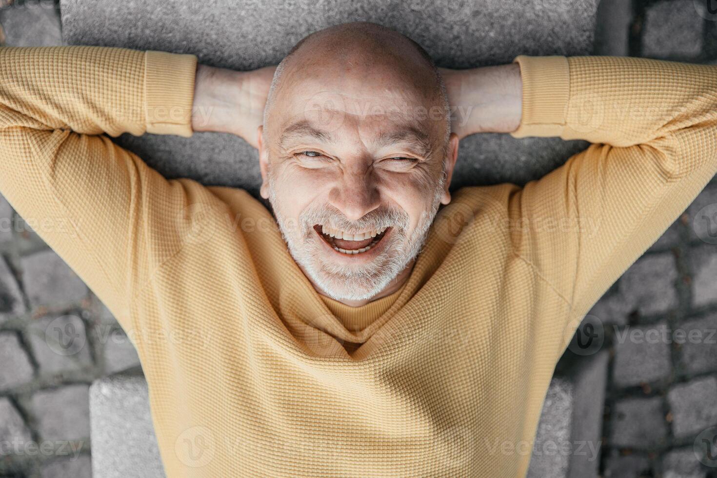 Playful bald senior man lying on his back on an urban bench, making funny faces, capturing a moment of levity photo