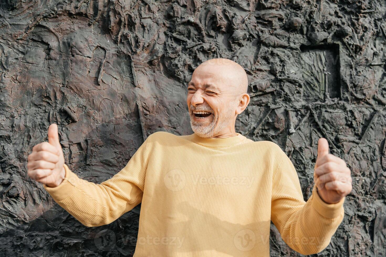 Joyful bald senior man in a yellow sweater giving thumbs up with a wide smile against a textured dark wall photo