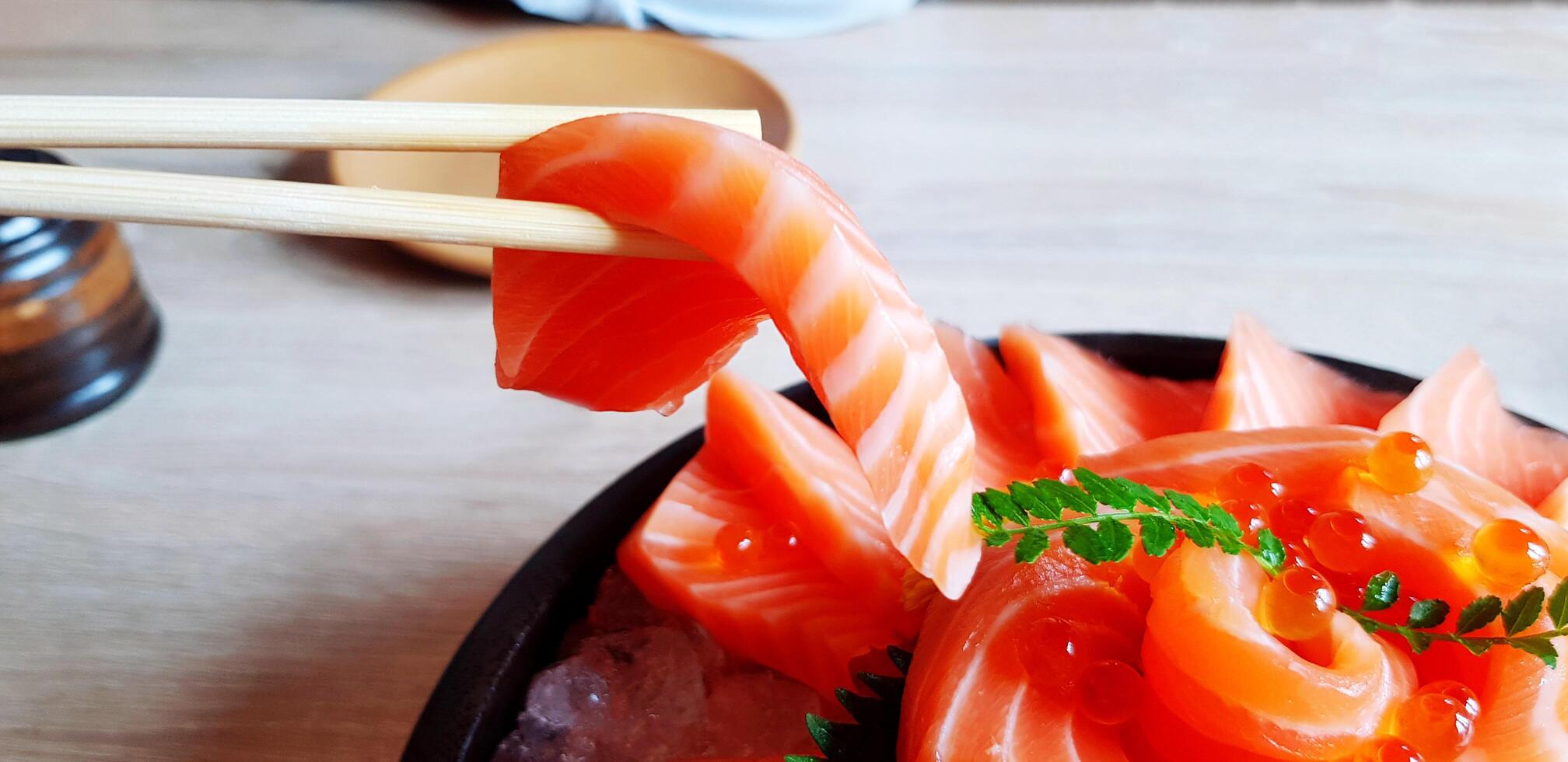 Close up eating fresh salmon by using chopsticks in black plate and on wooden bowl with copy space at Japanese restaurant. Sashimi or Healthy food, Diet and Life style eating . photo