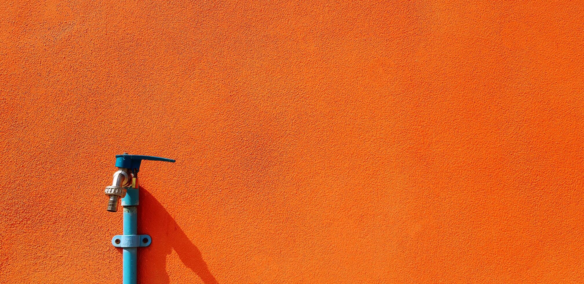 Blue faucet or tap and water pipe installed on orange concrete wall with right copy space. Object, colorful background and Public utility concept. photo