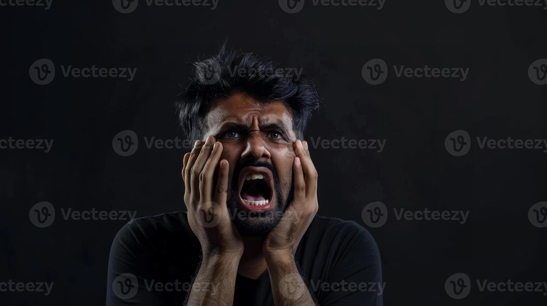 An Indian man with his hands on his face screaming in pain isolated against a black background photo